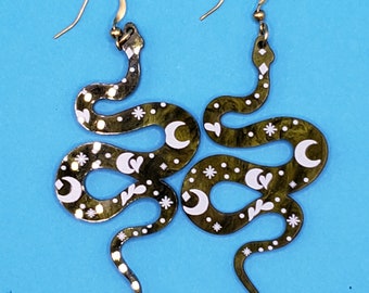 May include: A pair of gold snake earrings with white moon and star designs. The earrings are made of metal and have a dangling design.
