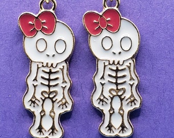 May include: A pair of dangle earrings featuring a white skeleton with a gold outline and a red bow on top. The earrings are on a purple background.