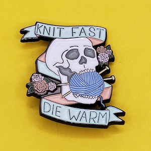 May include: Enamel pin featuring a skull with a blue yarn ball and knitting needles. The pin has a light blue banner with the words "KNIT FAST" and "DIE WARM". Pink and rose-colored flowers are on the sides.