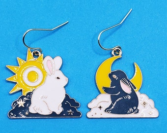 May include: A pair of gold-tone earrings featuring a white rabbit with a yellow sun and a black rabbit with a yellow moon. The earrings are designed with a cloud-like shape around the rabbits.