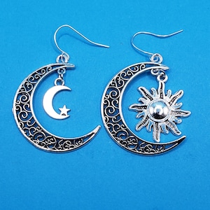 May include: A pair of silver-colored celestial-themed earrings. One earring features a crescent moon with a dangling star, the other a crescent moon with a sun pendant. Both crescent moons have a decorative filigree design. The earrings hang from silver-colored ear wires.