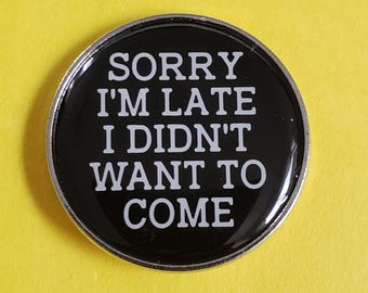 May include: A round, black pin with a silver border. The pin features the white text "SORRY I'M LATE I DIDN'T WANT TO COME." The pin is set against a yellow background.