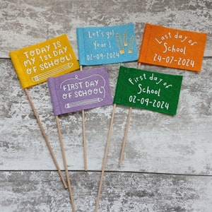 May include: Five felt flags with different messages about the first and last day of school. The flags are yellow, blue, purple, orange, and green. The flags are on wooden sticks. The text on the flags reads: "Today is my 1st day of school!" "Let's go! Year 1 02-09-2024" "First day of school" "Last day of school 24-07-2024" "First day of school 02-09-2024"