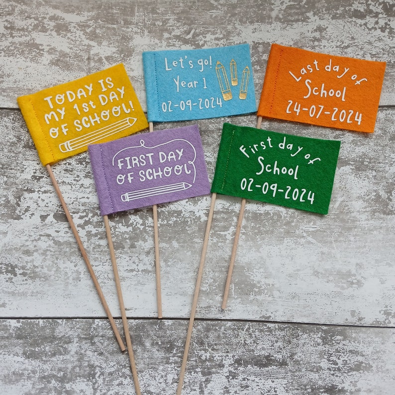 May include: Five felt flags with different messages about the first and last day of school. The flags are yellow, blue, purple, orange, and green. The flags are on wooden sticks. The text on the flags reads: "Today is my 1st day of school!" "Let's go! Year 1 02-09-2024" "First day of school" "Last day of school 24-07-2024" "First day of school 02-09-2024"