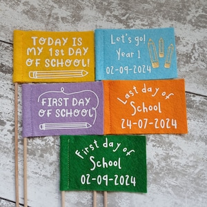 May include: Four felt flags with different messages about the first and last day of school. The flags are yellow, purple, blue, and green. The text on the flags reads: "Today is my 1st day of school!" "First day of school" "Let's go! Year 1 02-09-2024" "Last day of school 24-07-2024" "First day of school 02-09-2024"