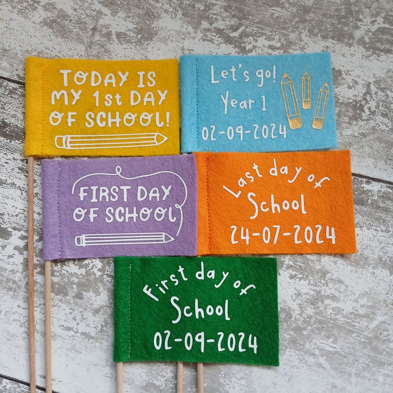 May include: Four felt flags with different messages about the first and last day of school. The flags are yellow, purple, blue, and green. The text on the flags reads: "Today is my 1st day of school!" "First day of school" "Let's go! Year 1 02-09-2024" "Last day of school 24-07-2024" "First day of school 02-09-2024"