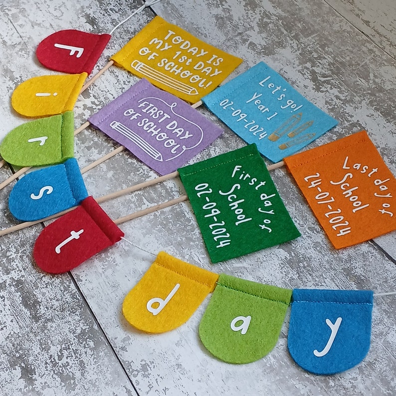 May include: A colorful felt banner with the words "First day of school" and the date "02-09-2024". The banner is made up of individual flags with the letters "First day" and the date on separate flags. The flags are attached to wooden sticks.