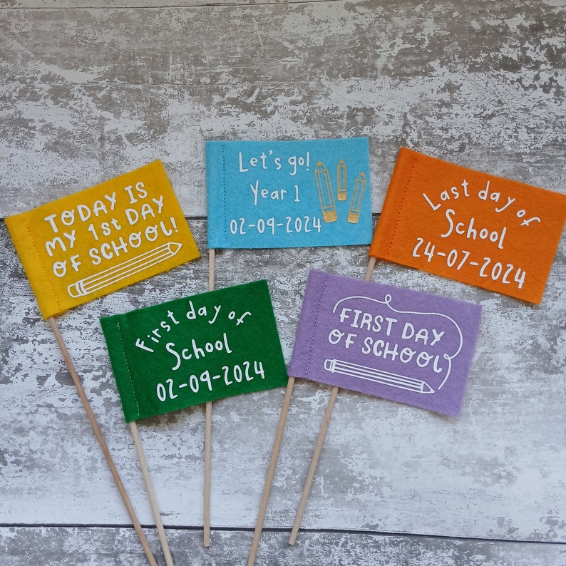 May include: Five felt flags with wooden sticks, each with a different message about the first and last day of school. The flags are yellow, blue, green, purple, and orange. The text on the flags reads: "Today is my 1st day of school!" "Let's go! Year 1 02-09-2024" "First day of school 02-09-2024" "Last day of school 24-07-2024" "First day of school"