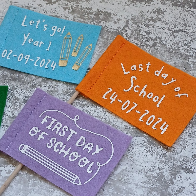 May include: Three felt flags with different messages for the first and last day of school. The first flag is blue with gold pencils and the text "Let's go! Year 1 02-09-2024". The second flag is purple with a white pencil and the text "FIRST DAY OF SCHOOL". The third flag is orange with white text that says "Last day of School 24-07-2024".
