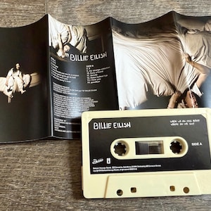 Op de afbeelding: Een cr&egrave;mekleurige cassetteband met zwarte labels met de tekst Billie Eilish en When We All Fall Asleep, Where Do We Go? samen met een bijpassende zwart-witte uitvouwbare j-card inlay met tracklijsten en fotografie.