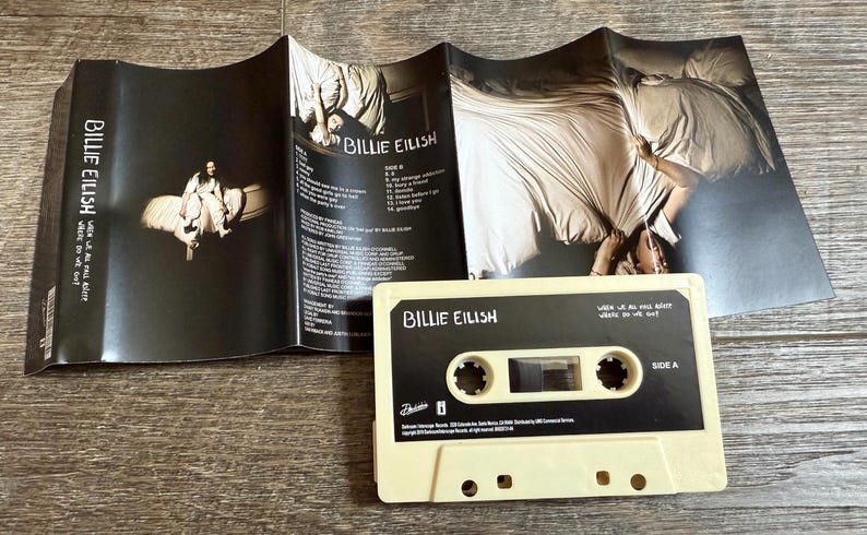 Op de afbeelding: Een cr&egrave;mekleurige cassetteband met zwarte labels met de tekst Billie Eilish en When We All Fall Asleep, Where Do We Go? samen met een bijpassende zwart-witte uitvouwbare j-card inlay met tracklijsten en fotografie.