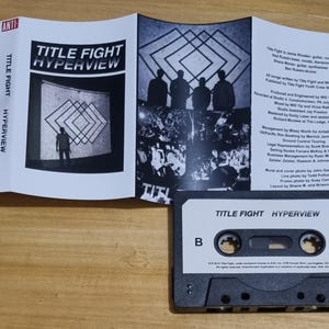 K&ouml;nnte beinhalten: Ein schwarzes Kassettenband mit dem Titel "TITLE FIGHT HYPERVIEW" auf dem Etikett. Das Band ist mit einer "B"-Seite beschriftet und hat ein "ANTI"-Logo auf der rechten Seite.