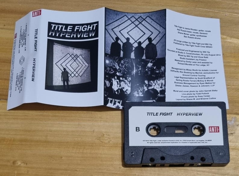 K&ouml;nnte beinhalten: Ein schwarzes Kassettenband mit dem Titel "TITLE FIGHT HYPERVIEW" auf dem Etikett. Das Band ist mit einer "B"-Seite beschriftet und hat ein "ANTI"-Logo auf der rechten Seite.