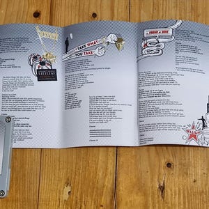 Może przedstawiać: Szara kaseta magnetofonowa z napisem "Lily Allen Alright, Still" leży przed rozłożoną wkładką albumu. Wkładka zawiera teksty piosenek i ilustracje na jasnoszarym tle. Kaseta magnetofonowa leży na drewnianej powierzchni.