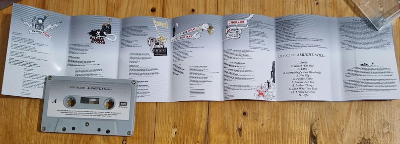 Może przedstawiać: Szara kaseta magnetofonowa z napisem "Lily Allen Alright, Still" leży przed rozłożoną wkładką albumu. Wkładka zawiera teksty piosenek i ilustracje na jasnoszarym tle. Kaseta magnetofonowa leży na drewnianej powierzchni.