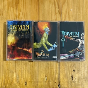 Può includere: Tre musicassette con copertine di album della band Trivium. Le musicassette sono in custodie di plastica trasparente. Le copertine degli album presentano illustrazioni con schemi di colori scuri, tra cui fiamme, una figura alata e un guerriero.