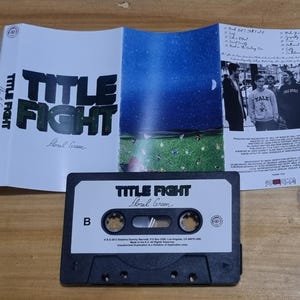 K&ouml;nnte beinhalten: Ein schwarzes Kassettenband mit dem Titel "Title Fight Floral Green" auf der Vorderseite. Das Kassettenband befindet sich in einem durchsichtigen Plastikgeh&auml;use mit einem wei&szlig;en Papiereinleger. Der Einleger zeigt auf der R&uuml;ckseite ein Schwarzwei&szlig;foto einer Band und auf der Vorderseite den Bandnamen und den Albumtitel.