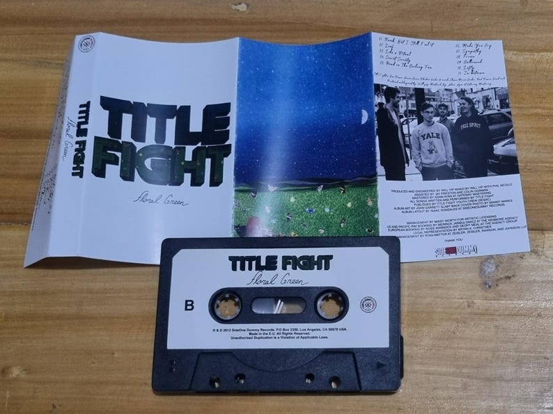 K&ouml;nnte beinhalten: Ein schwarzes Kassettenband mit dem Titel "Title Fight Floral Green" auf der Vorderseite. Das Kassettenband befindet sich in einem durchsichtigen Plastikgeh&auml;use mit einem wei&szlig;en Papiereinleger. Der Einleger zeigt auf der R&uuml;ckseite ein Schwarzwei&szlig;foto einer Band und auf der Vorderseite den Bandnamen und den Albumtitel.