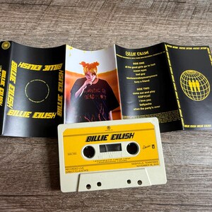 Op de afbeelding: Een Billie Eilish Live at Third Man Records cassettebandje met een witte behuizing en een geel label. De uitgevouwen inlay heeft een zwarte achtergrond met gele tekst, een foto van Billie Eilish en een tracklist voor kant &eacute;&eacute;n en twee.