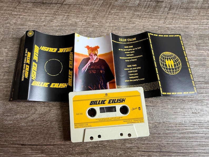 Op de afbeelding: Een Billie Eilish Live at Third Man Records cassettebandje met een witte behuizing en een geel label. De uitgevouwen inlay heeft een zwarte achtergrond met gele tekst, een foto van Billie Eilish en een tracklist voor kant &eacute;&eacute;n en twee.