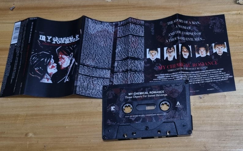 Puede incluir: Una cinta de casete negra con el t&iacute;tulo "My Chemical Romance Three Cheers For Sweet Revenge" impreso en la parte delantera. La cinta de casete est&aacute; en una caja de pl&aacute;stico con una imagen en blanco y negro de una banda en la parte delantera.