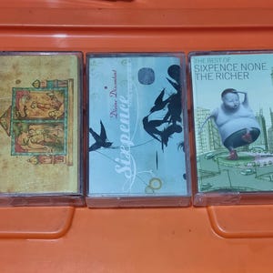 以下が含まれることがあります： 異なるアルバムカバーのついたカセットテープが3つあります。最初のテープは、薄い茶色のカバーに中世風のイラストが描かれています。2番目のテープは、薄い青色のカバーに白黒の鳥のイラストが描かれています。3番目のテープは、緑色のカバーに青いシャツと赤いズボンを着た人の漫画のイラストが描かれています。3番目のテープのテキストは「The Best of Sixpence None The Richer」です。