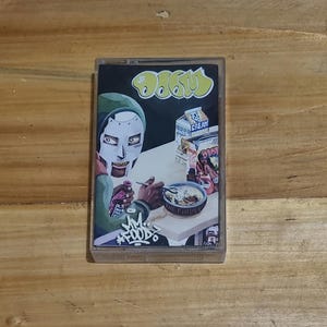 May include: A cassette tape with a black and green illustration of a masked figure eating cereal. The text "MF DOOM" is written in graffiti style on the bottom of the cassette tape.