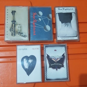 May include: Five cassette tapes featuring different Foo Fighters album covers. The covers display a range of designs, including a heart, a gun, a band logo, and a drawing of a person with a tattoo. The text on the tapes reads "Foo Fighters" and "There is nothing left to lose".