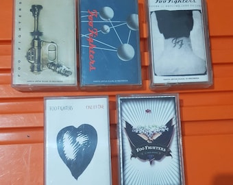 Puede incluir: Cinco cintas de cassette con diferentes portadas de álbumes de Foo Fighters. Las portadas presentan una variedad de diseños, que incluyen un corazón, un arma, un logotipo de la banda y un dibujo de una persona con un tatuaje. El texto en las cintas dice "Foo Fighters" y "There is nothing left to lose".