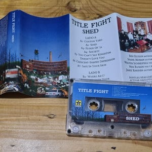 K&ouml;nnte beinhalten: Ein Kassettenband mit einem durchsichtigen Plastikgeh&auml;use. Das Band ist mit "Title Fight Shed" beschriftet. Das Kassettenband liegt auf einer Holzoberfl&auml;che. Das Band ist eine Musikaufnahme der Band Title Fight.