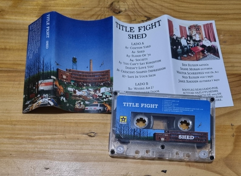 K&ouml;nnte beinhalten: Ein Kassettenband mit einem durchsichtigen Plastikgeh&auml;use. Das Band ist mit "Title Fight Shed" beschriftet. Das Kassettenband liegt auf einer Holzoberfl&auml;che. Das Band ist eine Musikaufnahme der Band Title Fight.
