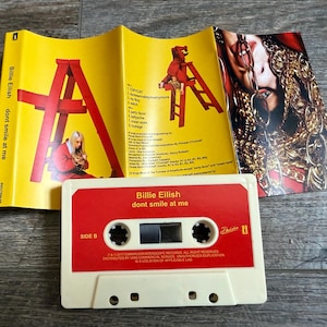 Op de afbeelding: Een Billie Eilish dont smile at me cassettebandje met een cr&egrave;mekleurige behuizing en een rood label. De bijbehorende gele J-card inlay toont een rode ladder, een persoon in een rode outfit en een foto van een persoon met gouden kettingen.