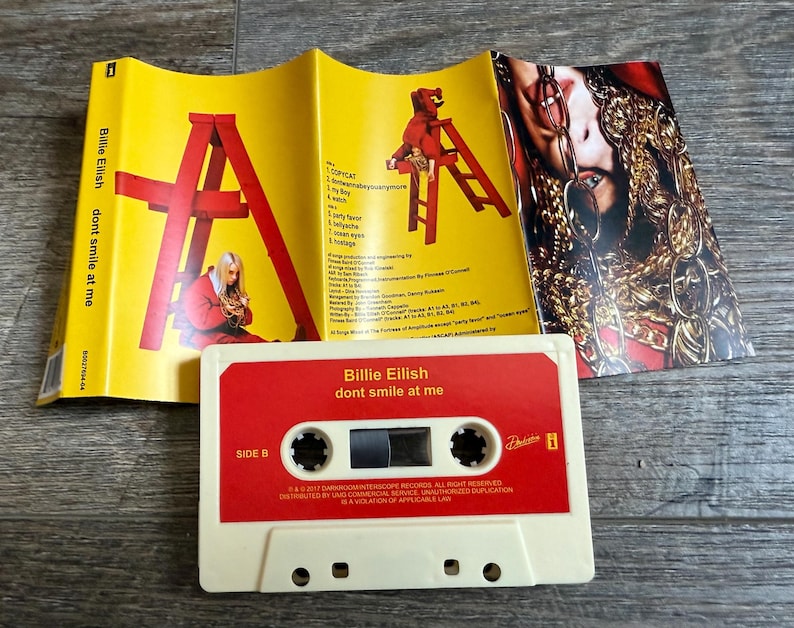 Op de afbeelding: Een Billie Eilish dont smile at me cassettebandje met een cr&egrave;mekleurige behuizing en een rood label. De bijbehorende gele J-card inlay toont een rode ladder, een persoon in een rode outfit en een foto van een persoon met gouden kettingen.