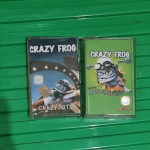 Könnte beinhalten: Zwei Kassetten mit grüner und gelber Verpackung, auf denen eine Comic-Frosch abgebildet ist. Die erste Kassette trägt den Titel "Crazy Frog Presents Crazy Hits". Die zweite Kassette trägt den Titel "Crazy Frog Presents More Crazy Hits".