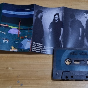 K&ouml;nnte beinhalten: Ein Kassettenband mit einem blau-schwarzen Geh&auml;use. Das Band ist mit "TITLE FIGHT" beschriftet und hat einen Bandnamen und einen Albumtitel, die auf dem Geh&auml;use aufgedruckt sind. Das Geh&auml;use zeigt ein Schwarzwei&szlig;bild einer Band auf der Vorderseite.