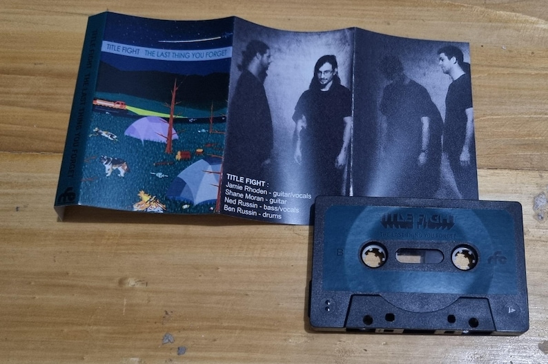 K&ouml;nnte beinhalten: Ein Kassettenband mit einem blau-schwarzen Geh&auml;use. Das Band ist mit "TITLE FIGHT" beschriftet und hat einen Bandnamen und einen Albumtitel, die auf dem Geh&auml;use aufgedruckt sind. Das Geh&auml;use zeigt ein Schwarzwei&szlig;bild einer Band auf der Vorderseite.