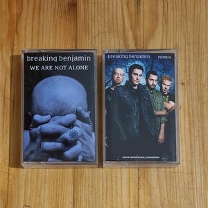 Może przedstawiać: Dwie kasety magnetofonowe, jedna z czarno-białym obrazkiem dłoni zasłaniającej twarz z tekstem "breaking benjamin WE ARE NOT ALONE", a druga z kolorowym obrazkiem zespołu z tekstem "breaking benjamin PHOBIA" i "HANYA UNTUK DIJUAL DI INDONESIA".