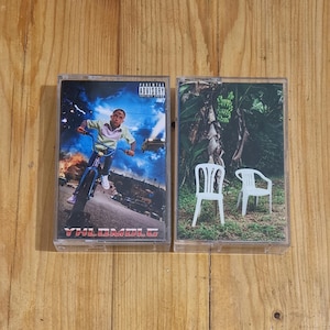 Può includere: Due cassette con copertine di album diverse. La prima cassetta ha una copertina blu e rossa con una persona che va in bicicletta e il testo "YHLQMDLG". La seconda cassetta ha una copertina verde con due sedie bianche e una pianta di banana.
