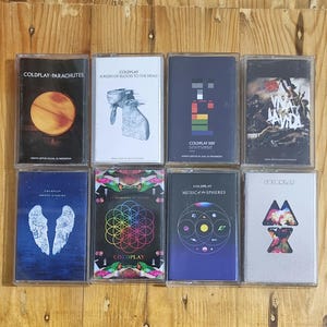 Può includere: Una collezione di otto cassette audio con la band Coldplay. Le cassette sono disposte in due file da quattro. Le copertine degli album presentano vari design, tra cui un pianeta, uno schema geometrico e un'immagine stilizzata di un cuore.