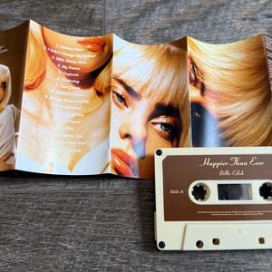 Op de afbeelding: Een Billie Eilish Happier Than Ever cassettebandje met de uitgevouwen inlay. De inlay bevat meerdere portretten van een blond persoon en een tracklist. Het cassettebandje is cr&egrave;mekleurig met een bruin label met de titel en artiest.