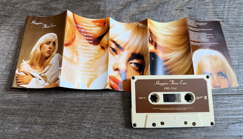 Op de afbeelding: Een Billie Eilish Happier Than Ever cassettebandje met de uitgevouwen inlay. De inlay bevat meerdere portretten van een blond persoon en een tracklist. Het cassettebandje is cr&egrave;mekleurig met een bruin label met de titel en artiest.