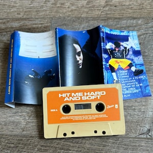 Op de afbeelding: Een beige cassettebandje met het label Hit Me Hard and Soft Side A, vergezeld door een blauw getinte J-card inlay met een persoon onder water, een close-up portret en een tracklist inclusief Skinny, Lunch en Blue met een persoon in een gele korte broek.