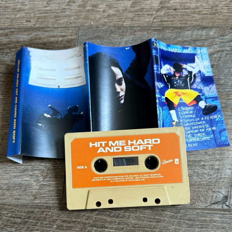Op de afbeelding: Een beige cassettebandje met het label Hit Me Hard and Soft Side A, vergezeld door een blauw getinte J-card inlay met een persoon onder water, een close-up portret en een tracklist inclusief Skinny, Lunch en Blue met een persoon in een gele korte broek.