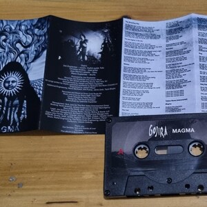 Pu&ograve; includere: Una cassetta audio nera con il testo "Gojira Magma" stampato sul davanti. La cassetta audio &egrave; su una superficie di legno. La cassetta audio fa parte di un set pi&ugrave; grande che include un CD e un libretto con i testi.