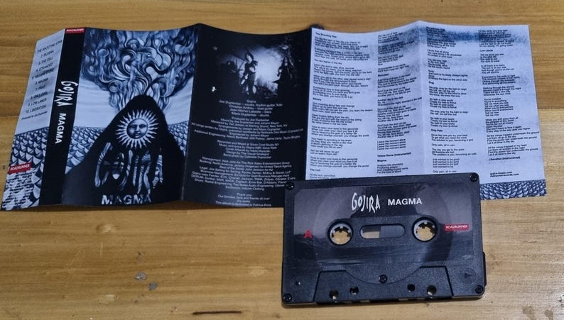 Pu&ograve; includere: Una cassetta audio nera con il testo "Gojira Magma" stampato sul davanti. La cassetta audio &egrave; su una superficie di legno. La cassetta audio fa parte di un set pi&ugrave; grande che include un CD e un libretto con i testi.