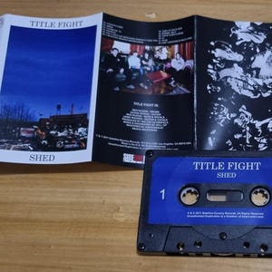 K&ouml;nnte beinhalten: Ein blaues Kassettenband mit dem Text "TITLE FIGHT SHED" auf der Vorderseite. Das Band ist mit der Nummer "1" beschriftet und hat ein schwarzes Kassettengeh&auml;use.