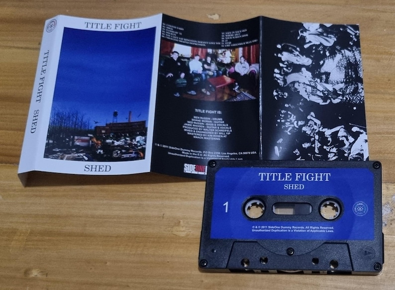 K&ouml;nnte beinhalten: Ein blaues Kassettenband mit dem Text "TITLE FIGHT SHED" auf der Vorderseite. Das Band ist mit der Nummer "1" beschriftet und hat ein schwarzes Kassettengeh&auml;use.