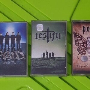 Pode incluir: Três fitas cassete com diferentes nomes de bandas e capas de álbuns. A primeira fita é uma imagem em preto e branco de uma banda chamada "P.O.D." com o título do álbum "Satellite". A segunda fita tem uma imagem em preto e branco de uma banda chamada "Testify" com uma cruz atrás do nome da banda. A terceira fita tem uma imagem em marrom e preto de uma banda chamada "P.O.D." com o título do álbum "Payable on Death" e uma borboleta com uma mulher no centro.