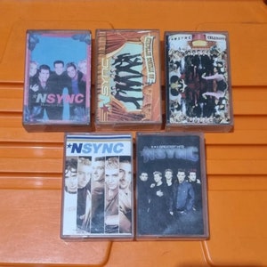 Op de afbeelding: Vijf cassettebandjes met de boyband *NSYNC. De bandjes zijn allemaal in doorzichtige plastic hoesjes met kleurrijke illustraties. De titels van de bandjes zijn "Home for Christmas", "No Strings Attached", "Celebrity", "Greatest Hits" en "NSYNC".