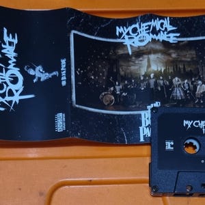 Puede incluir: Una cinta de casete negra con el texto "My Chemical Romance The Black Parade" en la parte delantera. La cinta de casete est&aacute; en una caja de pl&aacute;stico con una imagen en blanco y negro de la banda en la parte delantera.